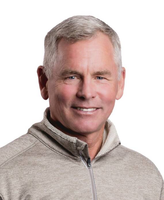 Portrait of a smiling gray-haired man in a beige zip-up sweater with a neutral background.