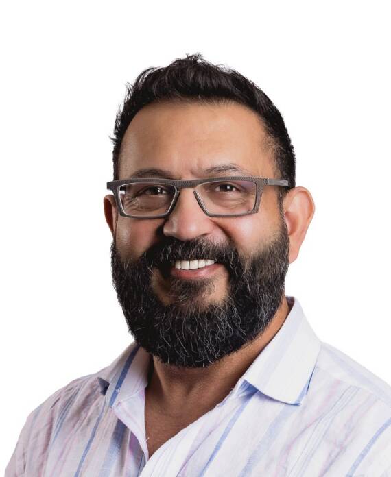 Portrait of a smiling man with a dark beard and glasses, wearing a light striped shirt, against a white background.