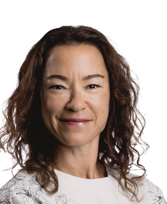 Smiling woman with shoulder-length curly brown hair wearing a light top, against a white background.