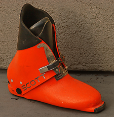 Bright orange rigid boot with a black padded ankle cuff and metal buckles, labeled SCOT, used for skiing or mountaineering gear