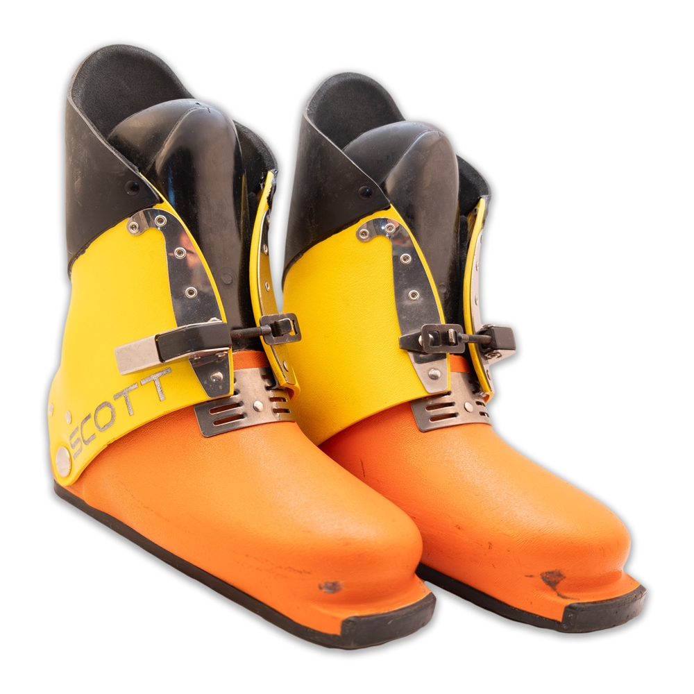 Pair of bright orange and yellow ski boots with black upper cuffs, buckles, and metal hardware shown on a white background.