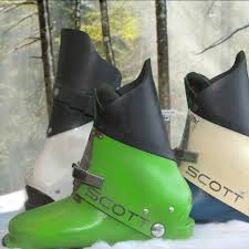 Green SCOTT ski boot in the foreground with two other boots (white and beige) behind, placed on snow in a forest setting.